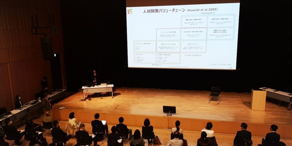 前方にスライドが投影され、カンファレンス参加者は真剣に講演を聞いています